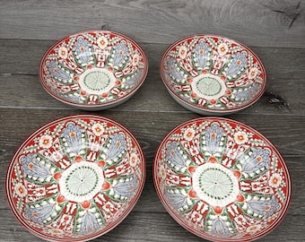 Pode incluir: Quatro tigelas de cerâmica decorativas com borda vermelha e intrincados padrões florais em azul, verde e branco. As tigelas têm um design circular no centro e estão dispostas sobre uma superfície de madeira cinza.