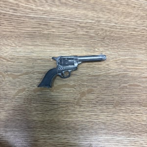 Op de afbeelding: Een klein, metalen speelgoedpistool met een donkergrijze handgreep en een zilveren loop. Het speelgoedpistool ligt op een houten oppervlak met een natuurlijk houtnerfpatroon. Het pistool is een miniatuur replica van een klassieke revolver.