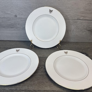 May include: Three white ceramic plates with a gold rim and a bee emblem. One plate is on a gold stand. The plates are oval and round. The plates are on a wooden surface.