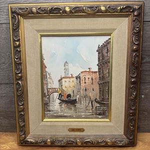 Könnte beinhalten: Ölgemälde einer venezianischen Kanalszene, gerahmt in einem goldenen, verzierten Rahmen. Das Kunstwerk zeigt Gebäude, eine Brücke und eine Gondel auf dem Wasser. Das Gemälde ist mit "DE VITY" signiert.