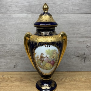 May include: A navy blue and gold decorative urn with a lid. The urn features gold handles and trim, and a central panel depicting a couple in a garden scene. The urn is approximately 30 cm tall.