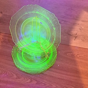 Op de afbeelding: Een stapel heldere neon-groene glazen borden met een bloemmotief. De borden hebben een geschulpte rand en worden gepresenteerd op een houten oppervlak. De borden zijn verlicht, waardoor een gloeiend effect ontstaat.