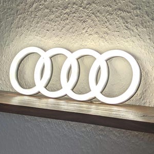 Może przedstawiać: Świecące białe logo Audi, składające się z czterech zazębiających się pierścieni, spoczywa na ciemnej drewnianej półce. Znak umieszczony jest na teksturowanej, kremowej ścianie. Pierścienie emitują miękkie światło.