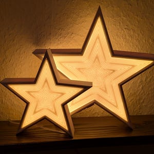 Könnte beinhalten: Zwei sternförmige Holzlampen mit warmem, leuchtendem Licht. Die Lampen haben ein geschichtetes Design mit einem dunkelbraunen Holzrahmen. Der größere Stern ist hinter dem kleineren positioniert, wodurch ein geschichteter Effekt entsteht. Die Lampen stehen auf einer Holzoberfläche.