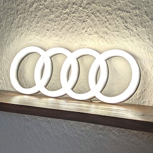 May include: A wall-mounted Audi logo sign featuring four interlocking rings illuminated with bright white LED light. The sign is positioned horizontally, casting a soft glow against the textured wall surface behind the light fixture.