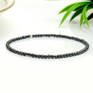 May include: A delicate, dark gray beaded bracelet. The bracelet is composed of small, faceted beads strung together, creating a flexible band. The beads have a subtle sparkle. The bracelet is displayed on a white surface with green foliage in the background.