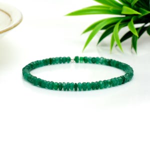Pode incluir: Uma delicada pulseira composta por pequenas contas de esmeralda facetadas. As contas são de uma cor verde vibrante e são enfiadas com um fecho prateado. A pulseira é exibida sobre uma superfície branca com folhagem verde no fundo.