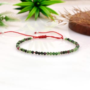 May include: A delicate bracelet with small, round green and red beads strung on a red cord. The bracelet has a sliding knot closure and silver-coloured accents. The beads are evenly spaced, creating a simple yet elegant design.