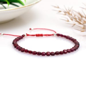 Op de afbeelding: Een delicate armband met kleine, gefacetteerde, dieprode edelstenen geregen op een rood koord. De verstelbare armband heeft een schuifknoopsluiting en zilverkleurige accenten. De sieraden worden gepresenteerd op een wit oppervlak.