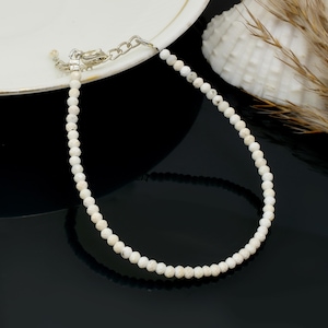May include: A delicate white beaded bracelet with a silver clasp and chain. The bracelet is composed of small, round, off-white beads. The jewelry is displayed on a black surface with a white plate and seashell in the background.
