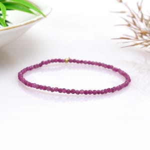 May include: A delicate bracelet featuring small, faceted, deep pink gemstones strung together. A small gold-colored bead is centered on the bracelet. The bracelet is displayed on a white surface with a blurred background.