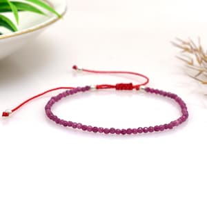 Op de afbeelding: Een delicate armband met kleine, gefacetteerde, magenta-kleurige edelstenen geregen op een rood koord. De armband heeft een schuifknoopsluiting voor verstelbare maatvoering en zilverkleurige accenten. De edelstenen zijn gelijkmatig verdeeld.