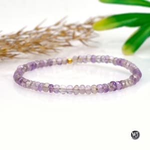 May include: A delicate bracelet composed of small, round, light purple amethyst beads. The beads are strung together, forming a complete circle. A small gold-colored bead is visible. The bracelet is displayed on a white surface with a blurred background.