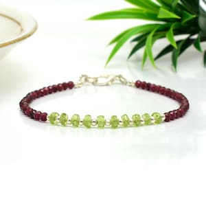 Könnte beinhalten: Ein filigranes Armband mit abwechselnden Abschnitten aus kleinen, runden, tiefroten Perlen und hellgrünen, unregelmäßig geformten Perlen. Das Armband wird mit einem silbernen Verschluss gesichert und auf einer weißen Oberfläche präsentiert.
