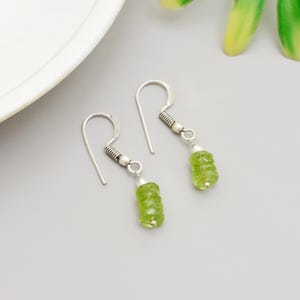 Può includere: Orecchini pendenti in argento con pietre preziose cilindriche verde chiaro. Gli orecchini hanno un semplice design a gancio con un accento a spirale vicino alla parte superiore. Le pietre preziose sono di un verde traslucido vibrante.