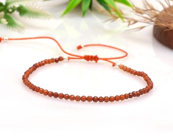 May include: Ankle bracelet featuring small, faceted, reddish-brown beads strung on an orange cord. The cord has silver-colored accents and a sliding knot closure. The jewelry is displayed on a white surface with green foliage in the background.