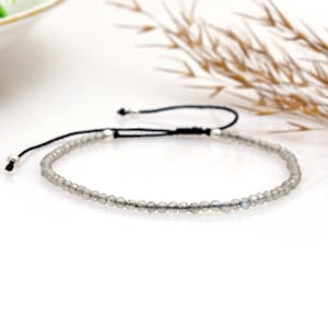 Puede incluir: Una delicada pulsera con pequeñas cuentas grises facetadas ensartadas en un cordón negro. La pulsera ajustable tiene extremos plateados y un cierre de nudo corredizo. Las cuentas parecen ser de un material translúcido.