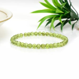 May include: A bracelet made of small, round, light green peridot beads. The beads are strung together, creating a circular shape. The bracelet is shown on a white surface, with green foliage in the background. This piece of jewellery is a perfect accessory.