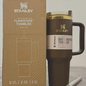 May include: A brown Stanley Flowstate tumbler with a gold lid and handle, alongside its cardboard packaging. The box and tumbler display the Stanley logo and product information, including "40 OZ | 1.18L". The tumbler is designed for all-day hydration.