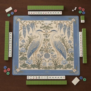 May include: A square mahjong mat featuring a central illustration of two herons facing each other amidst wisteria and lotus flowers, framed by a blue border. The mat is surrounded by rows of green and white mahjong tiles and colorful game chips.