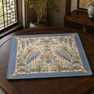 May include: A rectangular placemat featuring a cream-colored center with two blue and white herons facing each other, surrounded by floral motifs and wisteria. The border is a solid blue with geometric corner accents and a central circular emblem.