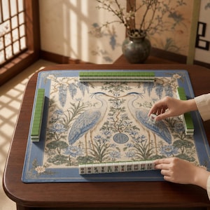 May include: A square mahjong mat featuring a cream-colored center with two blue and white herons surrounded by floral motifs, framed by a blue border. The mat is set on a wooden table with green mahjong tiles arranged in rows along the edges.