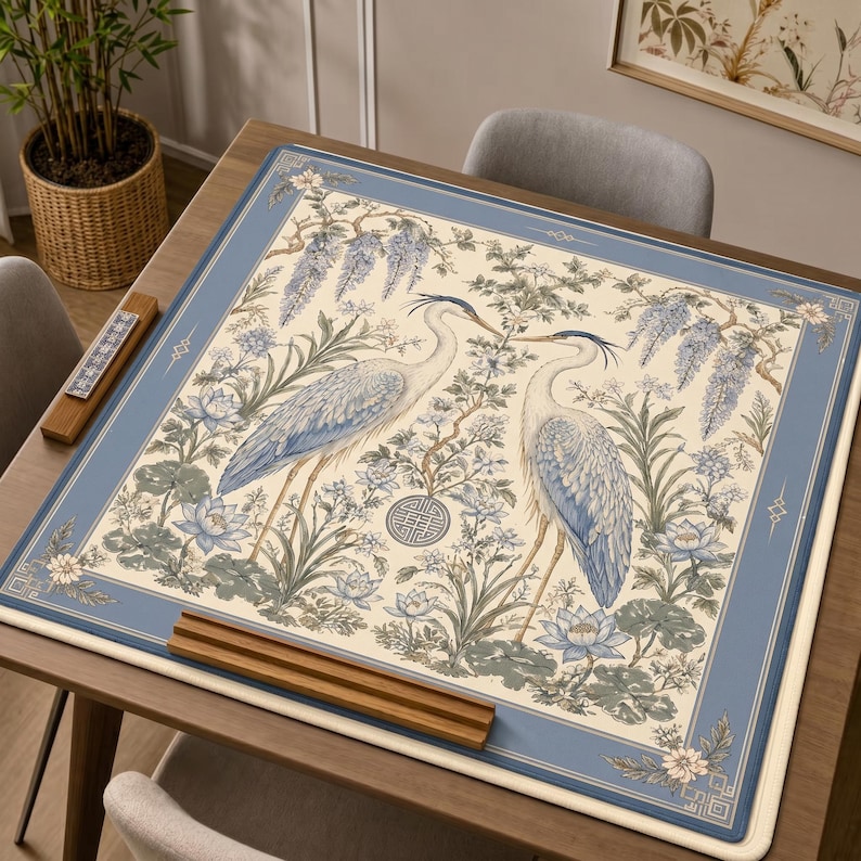 May include: A square table mat featuring a cream-colored center with two blue and white herons standing among lotus flowers and wisteria branches. The design includes a circular geometric emblem and a wide blue border with decorative corner accents.