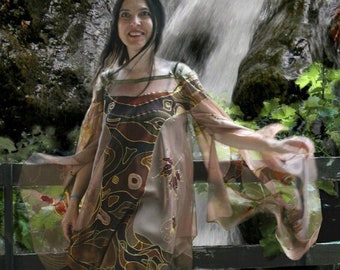 Puede incluir: Una mujer con una túnica estampada y una falda negra está de pie junto a una cascada. La túnica presenta diseños abstractos marrones y dorados. Lleva sandalias doradas. El fondo incluye una barandilla de madera y vegetación.