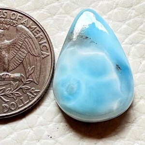 May include: A polished, teardrop-shaped gemstone with a light blue and white swirling pattern. A U.S. quarter coin is next to the gemstone for size comparison. The gemstone has a glossy finish and is set against a neutral background.