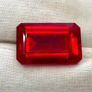 Może przedstawiać: Fasetowany, prostokątny, głęboko czerwony kamień szlachetny. Klejnot ma wypolerowaną powierzchnię i jest trzymany na białym tle z tkaniny. Kształt klejnotu to szlif szmaragdowy, z prostokątnymi fasetami.