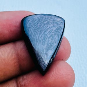 May include: A polished, black gemstone with a unique, swirling pattern. The triangular shape is held in a hand, showcasing the stone's smooth, reflective surface. The stone's surface has a light blue hue.