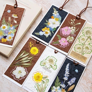 Könnte beinhalten: Eine Sammlung handgefertigter Lesezeichen mit gepressten Blumen. Jedes Lesezeichen hat ein einzigartiges Design mit einer Vielzahl farbenfroher Blumen und Grünpflanzen, vor einem braunen oder weißen Hintergrund. Sie haben eine Lederschnur zur einfachen Verwendung.