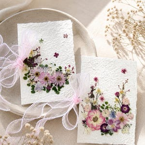 Pode incluir: Dois cartões retangulares de papel branco texturizado feito à mão, decorados com arranjos de flores secas prensadas em tons de rosa, roxo e verde. Cada cartão é amarrado com uma fita rosa claro transparente com bordas de miçangas.