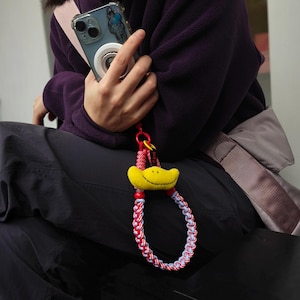 Könnte beinhalten: Ein gelber Handy-Anhänger in Entenform mit einer rot-weiß-blauen geflochtenen Handschlaufe. Der Anhänger hat ein lächelndes Gesicht und ist an einer Handyhülle mit einem silbernen Ringhalter befestigt.
