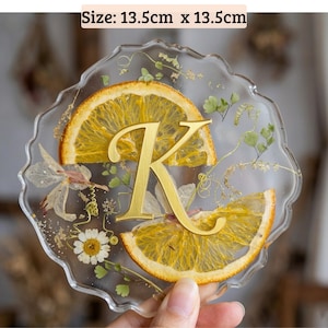 May include: A clear resin coaster, 13.5cm x 13.5cm, with dried orange slices, small white flowers, and gold accents. A gold letter "K" is centered on the coaster. The coaster has a wavy edge.