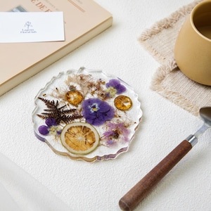 Puede incluir: Un posavasos de resina transparente con flores secas y rodajas de cítricos. El posavasos presenta un pensamiento morado, helechos secos y rodajas de naranja. Se ve una tarjeta de visita con un logotipo floral.