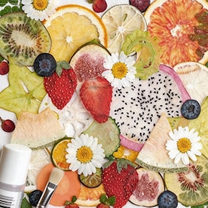 Puede incluir: Un vibrante arreglo de rodajas de fruta deshidratada, incluyendo kiwi, naranja y fruta del dragón, intercaladas con fresas frescas, arándanos y margaritas. Una pequeña botella y un pincel se encuentran en la esquina inferior izquierda.