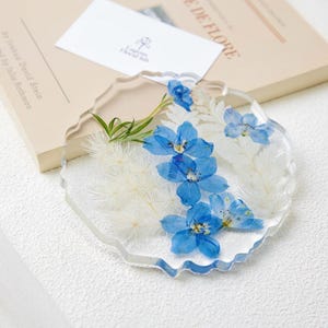Puede incluir: Un posavasos de resina transparente con borde ondulado, con flores azules de delfinio y detalles blancos plumosos. Una pequeña tarjeta blanca con un logotipo y un libro en el fondo. El posavasos tiene aproximadamente 10 cm de diámetro.