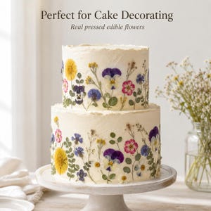 May include: A two-tiered cake with white frosting decorated with real pressed edible flowers in shades of yellow, purple, pink, and blue. The text "Perfect for Cake Decorating" and "Real pressed edible flowers" appears above the cake.