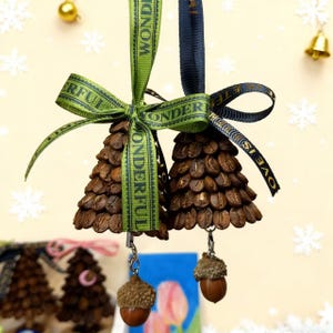 May include: Two Christmas tree ornaments crafted from coffee beans, adorned with green and navy blue ribbons inscribed with "Wonderful" and "Love is". Each ornament features a small acorn charm. The background is a pale yellow with snowflake details.