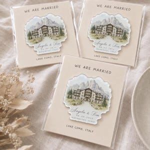 Puede incluir: Tres recuerdos de boda idénticos en fundas de plástico transparente, cada uno con un adorno acrílico festoneado con una ilustración en acuarela de un hotel de montaña. Texto: WE ARE MARRIED, Angela & Tim, 06.09.2026, DOLOMITES, ITALY, LAKE COMO, ITALY.