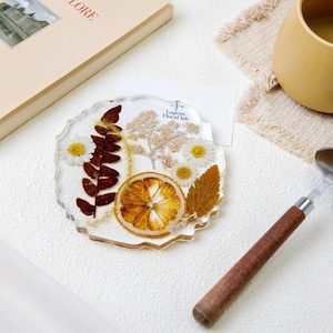 Puede incluir: Un posavasos de resina transparente con borde festoneado, con flores secas, hojas y una rodaja de naranja seca. El posavasos tiene el texto "Lagom Floral lab". Un artículo decorativo y único.