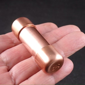May include: A copper-colored cylindrical object with rounded ends, held in a hand. The object has a smooth, metallic finish and appears to be a small container or component. The letters "BPC" are stamped on one end.