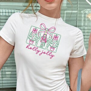 Pu&ograve; includere: Una t-shirt bianca a maniche corte con una stampa grafica di due alberi di Natale e uno schiaccianoci legati insieme da un fiocco rosa. Il testo "holly jolly" &egrave; stampato in corsivo rosa sotto l'illustrazione festiva natalizia.