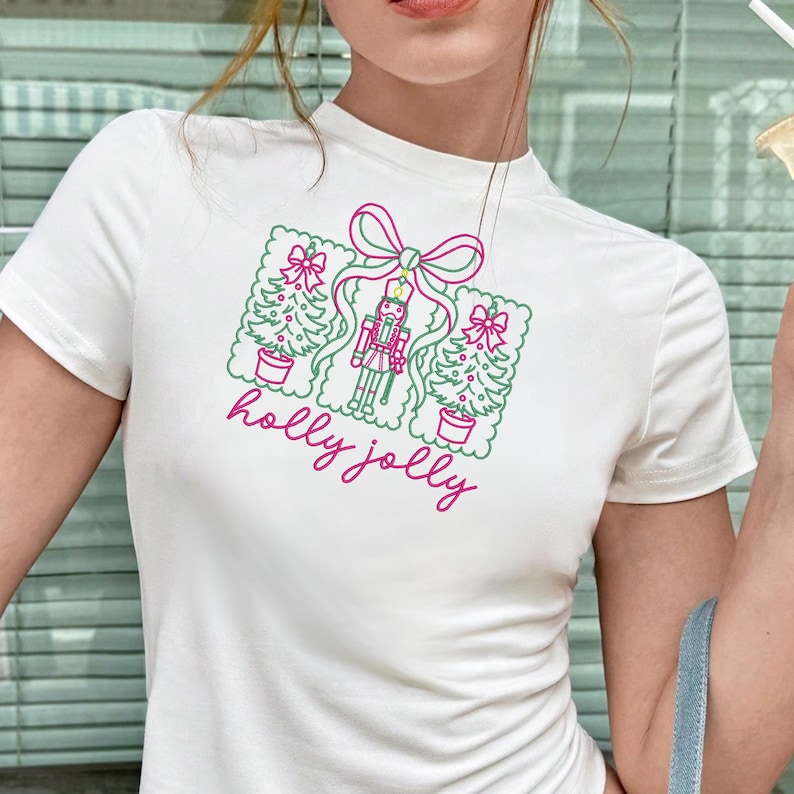 Pu&ograve; includere: Una t-shirt bianca a maniche corte con una stampa grafica di due alberi di Natale e uno schiaccianoci legati insieme da un fiocco rosa. Il testo "holly jolly" &egrave; stampato in corsivo rosa sotto l'illustrazione festiva natalizia.