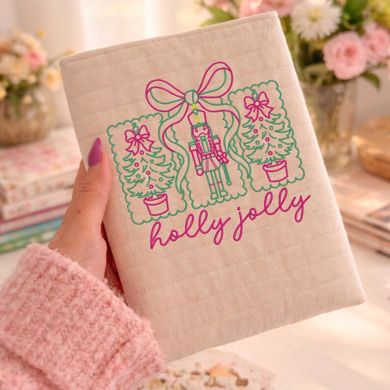 Pu&ograve; includere: Una copertina per libro in tessuto beige trapuntato con disegni ricamati in filo rosa e verde. Il ricamo raffigura uno schiaccianoci centrale affiancato da due alberi di Natale, il tutto legato da un grande fiocco. Il testo holly jolly &egrave; ricamato in rosa sotto i disegni.