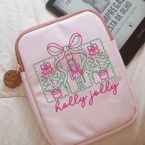 Pu&ograve; includere: Una custodia protettiva rosa chiaro con un ricamo raffigurante uno schiaccianoci, due alberi di Natale e un grande fiocco in filo verde e rosa. Il testo "holly jolly" &egrave; ricamato in corsivo rosa sotto le illustrazioni festive.