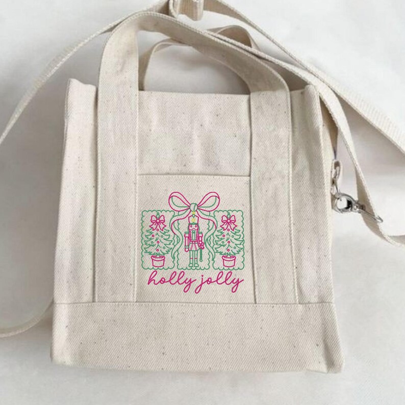 Pu&ograve; includere: Una borsa tote in tela color naturale con tracolla, caratterizzata da un ricamo rosa e verde raffigurante uno schiaccianoci affiancato da due alberi di Natale sotto un fiocco, con la scritta "holly jolly" in corsivo rosa sotto.