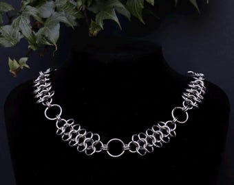 May include: A silver-colored chainmail necklace displayed on a black mannequin. The necklace features multiple rows of small, circular links connected to larger rings. The background is black with green foliage at the top.