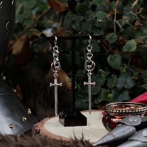 Può includere: Un paio di orecchini a forma di spada color argento appesi a un espositore nero. Gli orecchini presentano un ciondolo a forma di spada con elsa e lama dettagliate, collegato a una catena di piccoli anelli. Lo sfondo è un misto sfocato di fogliame verde.
